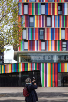 Béal & Blanckaert | Logements | La Courneuve Femme prenant en photo un bâtiment multicolore au marché des quatre chemins